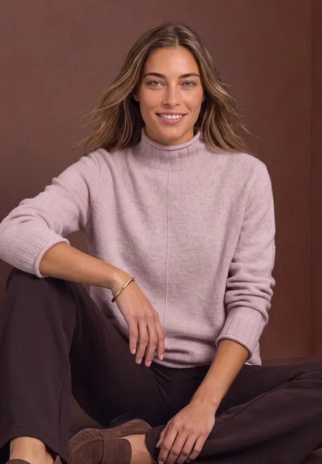 Une femme souriante est assise détendue, portant un pull rose et un pantalon marron devant un fond neutre.
