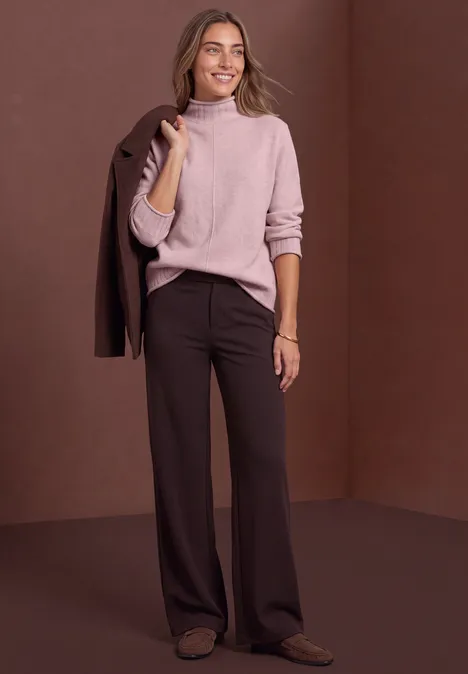 Femme élégante en pull rose et pantalon large marron, veste reposant sur l'épaule, souriante.