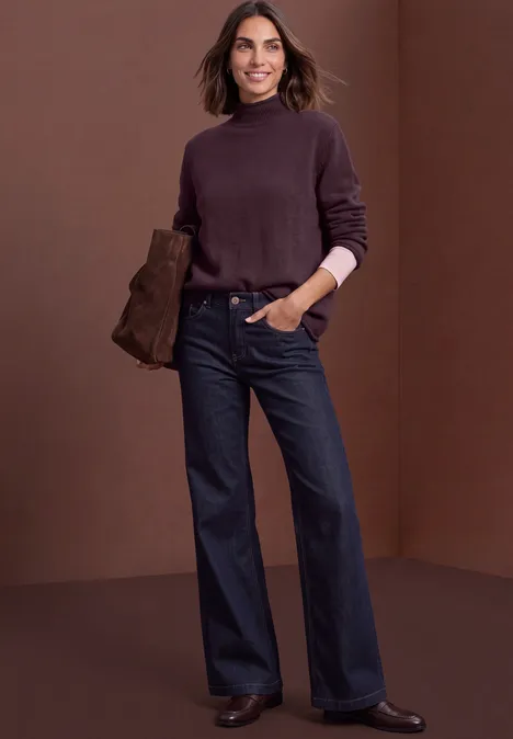 Vrouw in donkerpaarse coltrui en wijde jeans met clutch, poseert voor een bruine achtergrond.