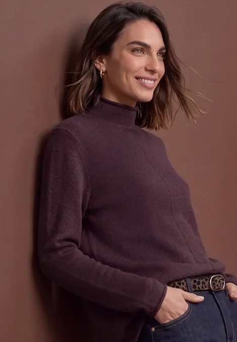 Een glimlachende vrouw in een bruin coltrui en jeans, tegen een muur geleund.