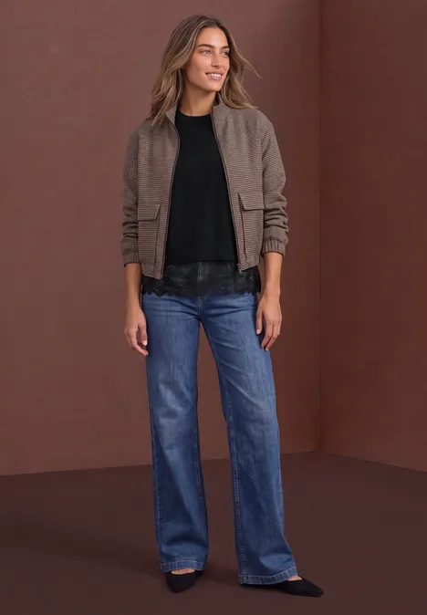 Frau in brauner Jacke, schwarzem Shirt und weiten Jeans vor braunem Hintergrund.
