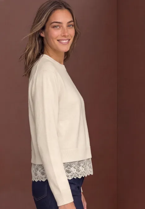 Frau in beigem Pullover mit Spitzenabschluss und Jeans, lächelnd vor braunem Hintergrund.