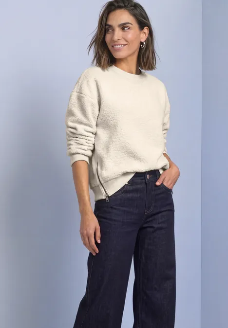 Eine Frau trägt einen hellen Pullover und dunkle Jeans vor neutralem Hintergrund.