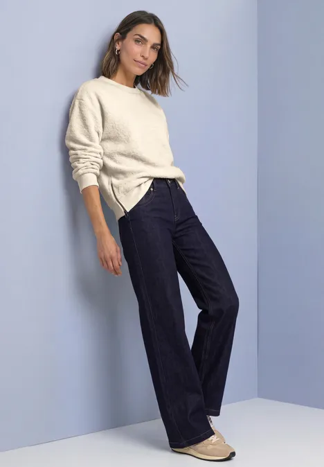 Frau lehnt an Wand, trägt cremefarbenen Pullover, dunkle Schlagjeans und helle Ballerinas vor schlichtem Hintergrund.
