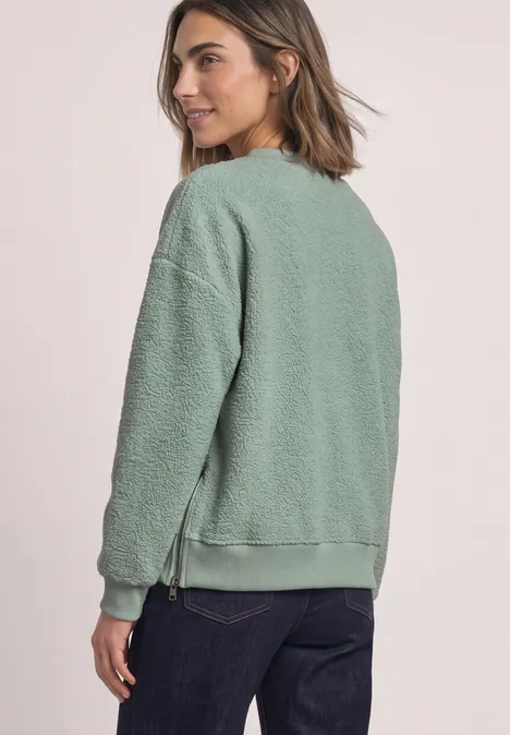 Seitliche Ansicht einer Frau in grünem Teddy-Fleece-Sweatshirt und dunklen Jeans vor hellem Hintergrund.