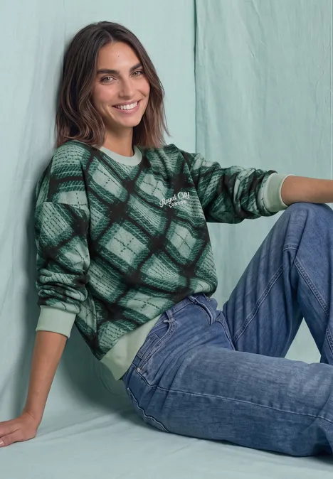 Eine lächelnde Frau sitzt vor einem pastellgrünen Hintergrund und trägt einen karierten grünen Pullover und Jeans.