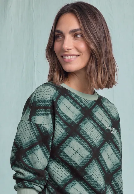 Eine lächelnde Frau im grünen karierten Pullover vor hellem Hintergrund.