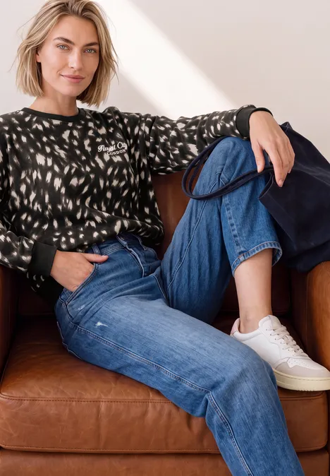 Sweatshirt mit Rundhals und Rehprint Black Eine Frau sitzt entspannt auf einem braunen Ledersofa, trägt ein gemustertes Sweatshirt, blaue Jeans und weiße Sneaker.