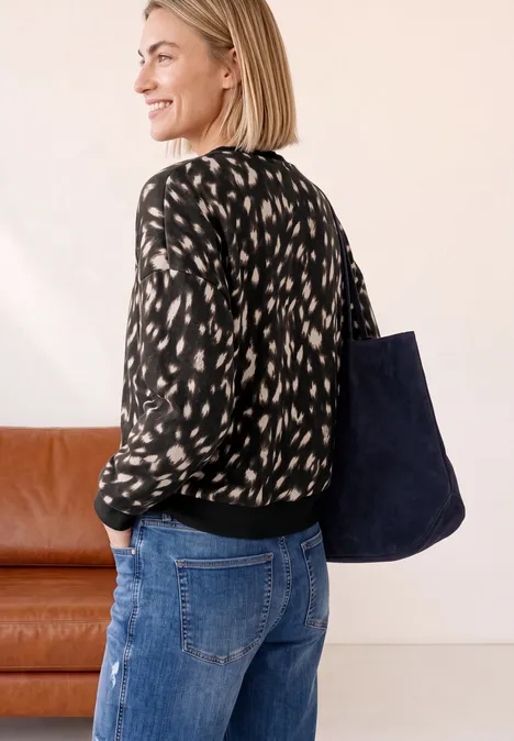 Sweatshirt mit Rundhals und Rehprint Black Frau in gemusterter Bluse und Jeans trägt eine große blaue Tasche, neben einem braunen Sofa.