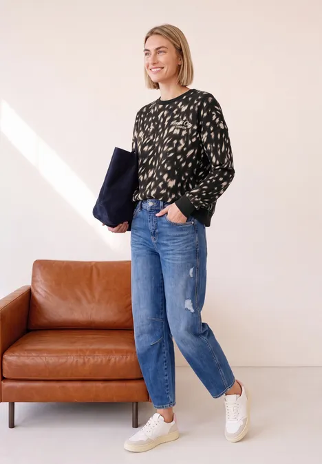 Sweatshirt mit Rundhals und Rehprint Black Frau mit schwarzem gemustertem Pullover, blauen Jeans und weißen Sneakern steht neben einem braunen Ledersofa.