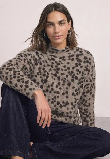Frau in beige-schwarzem Fleckenpullover und dunklen Jeans, sitzend vor einem neutralen Hintergrund.