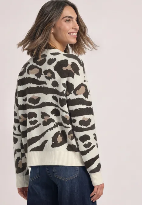 Vrouw in spijkerbroek en beige-zwart dierenprint trui, glimlachend opzij kijkend.