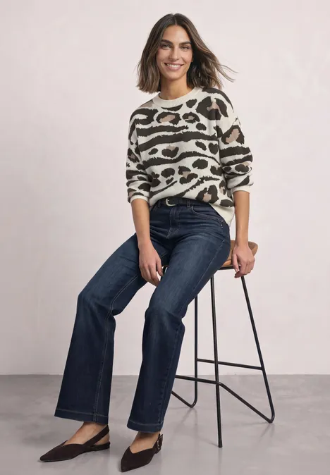 Vrouw zit op kruk in animalprint trui, rechte donkerblauwe jeans en zwarte pumps, ze lacht.