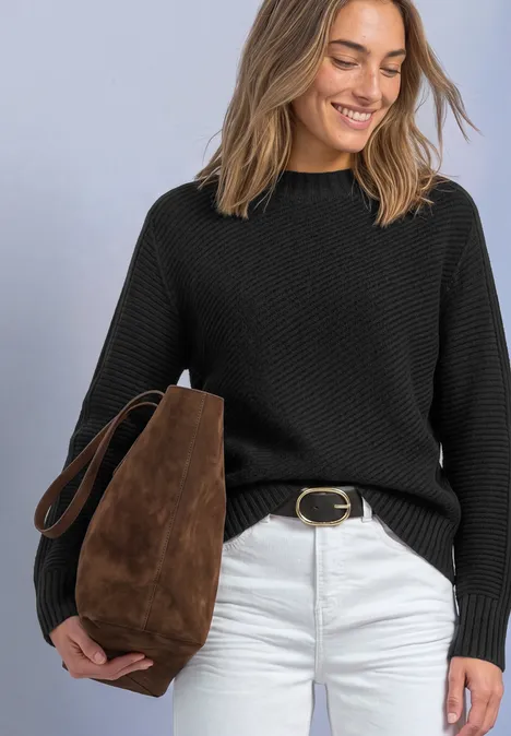 Frau mit schwarzem Strickpullover, weißer Hose und brauner Suede-Tasche, lächelt und blickt nach unten.