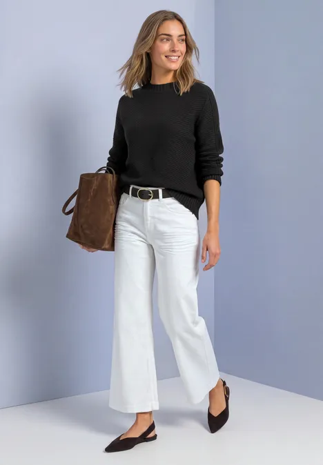 Frau in schwarzem Pullover und weißen, weiten Jeans trägt schwarze Pumps und eine braune Tasche vor hellem Hintergrund.