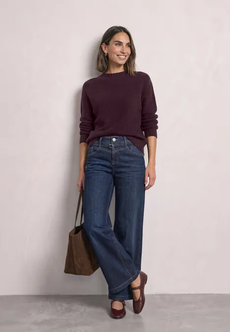Frau in lila Pullover, weitem Jeans und braunen Loafers mit passender Tasche.