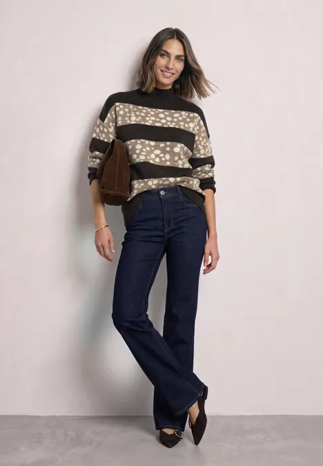 Vrouw in gestreepte, gevlekte trui, donkere jeans, pumps en bruine tas voor lichte achtergrond.