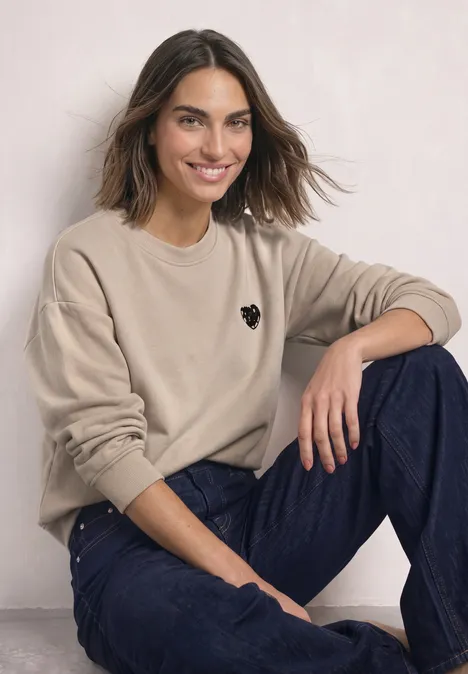 Loose Fit Sweatshirt mit Patch Detail clay sand Junge Frau sitzt lässig, trägt beigefarbenes Sweatshirt mit Herzlogo und blaue Jeans, lächelt entspannt.