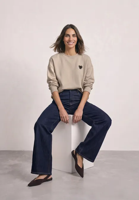 Loose Fit Sweatshirt mit Patch Detail clay sand Frau mit beiger Sweatshirt und weiten Jeans sitzt lächelnd auf einem weißen Hocker vor hellem Hintergrund.