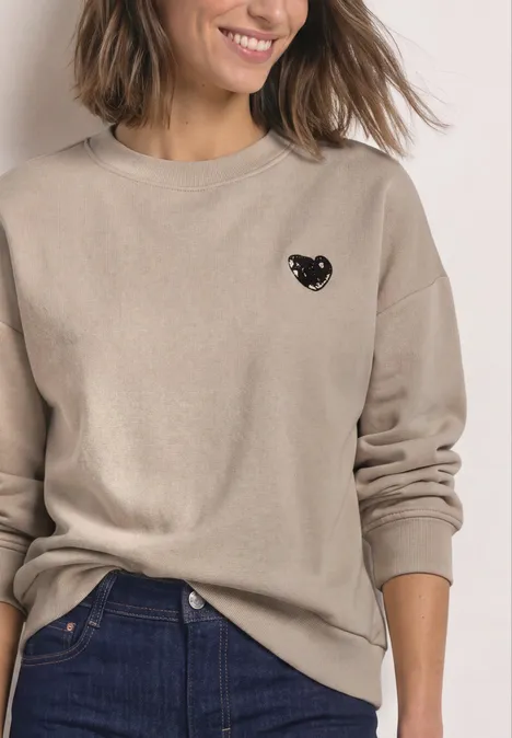 Loose Fit Sweatshirt mit Patch Detail clay sand Eine Frau trägt einen beigen Pullover mit kleinem schwarzem Katzen-Motiv und eine Jeans.