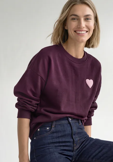 Loose Fit Sweatshirt mit Patch Detail berry maroon Lächelnde Frau in lila Sweatshirt mit Herzmotiv und blauen Jeans vor hellem Hintergrund.