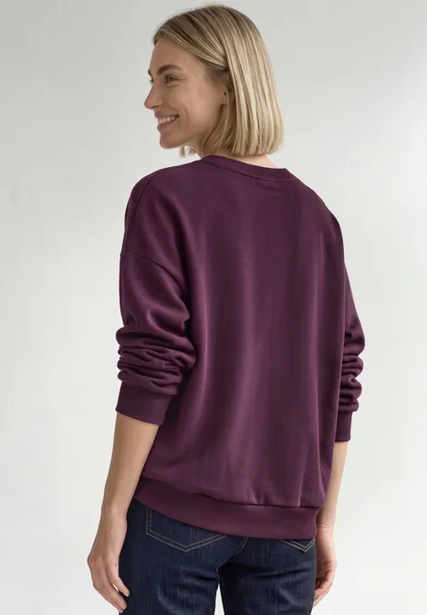 Loose Fit Sweatshirt mit Patch Detail berry maroon Frau von hinten mit hellblondem Bob, die ein violettes Sweatshirt trägt und lächelnd seitlich schaut.