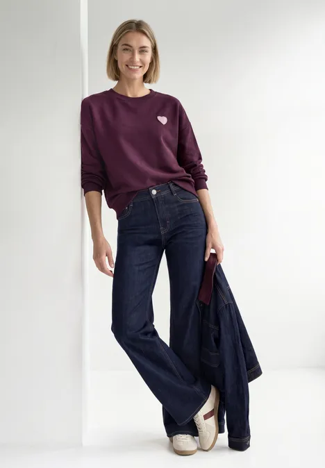 Loose Fit Sweatshirt mit Patch Detail berry maroon Frau in lila Pullover, weiter Jeans, weißen Sandalen, hält Jeansjacke und lächelt.