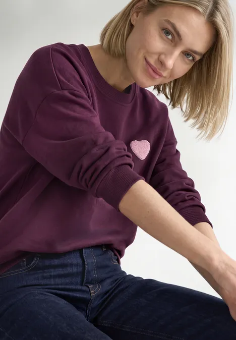 Loose Fit Sweatshirt mit Patch Detail berry maroon Eine lächelnde Frau in lila Sweatshirt mit Herzmotiv und blauen Jeans vor neutralem Hintergrund.
