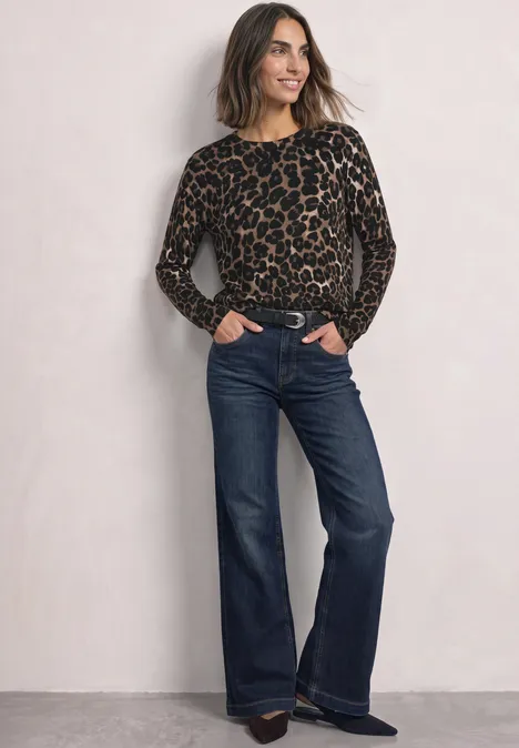 Frau in Leopard-Pulli und dunkelblauen, weiten Jeans, lächelt mit Händen in den Taschen vor neutralem Hintergrund.