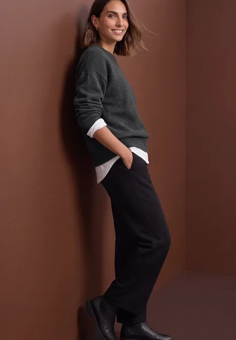 Pullover in Unifarbe mit Rundhals armor grey mel. Eine lächelnde Frau in grauem Pullover, weißem Shirt und schwarzen Hosen lehnt entspannt an einer braunen Wand.