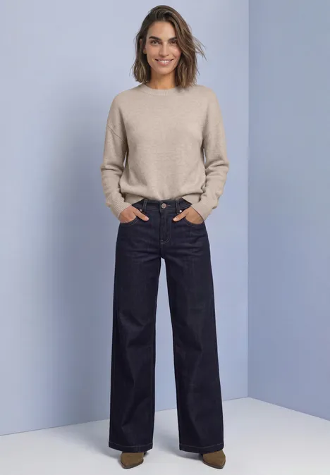 Frau mit beigem Pullover und weit geschnittenen dunklen Jeans, Hände in den Taschen, vor hellblauem Hintergrund.