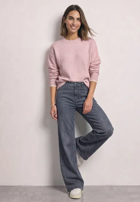 Frau in rosa Pullover und weiten Jeans vor neutralem Hintergrund.