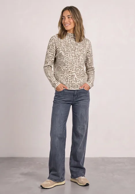 Pullover mit Turtle Neck und Jacquardmuster pure beige Frau in Pullover mit Tierprint, blauen Jeans und Sneakers steht entspannt vor neutralem Hintergrund.