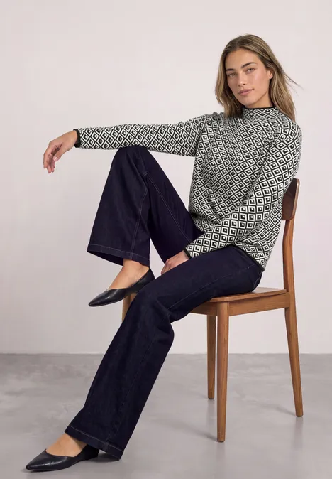 Pullover mit Turtle Neck und Jacquardmuster Black Frau sitzt auf Holzstuhl, trägt gepunktetes Oberteil, dunkle Jeans und schwarze Ballerinas.
