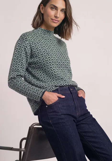 Pullover mit Turtle Neck und Jacquardmuster stormy green Frau in gemustertem Pullover und Jeans lehnt an einem Stuhl.