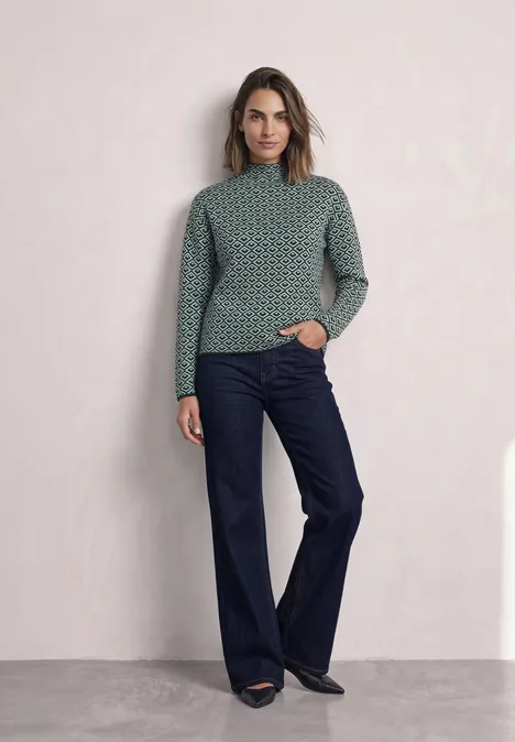 Pullover mit Turtle Neck und Jacquardmuster stormy green Frau in grün gemustertem Pullover und weiten dunkelblauen Jeans vor hellem Hintergrund.