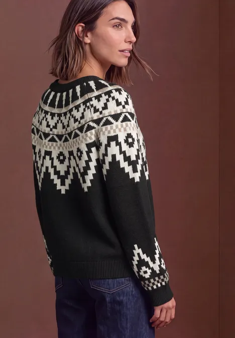 Eine Frau trägt einen schwarz-weißen Norwegerpullover und Jeans, schaut seitlich, vor braunem Hintergrund.