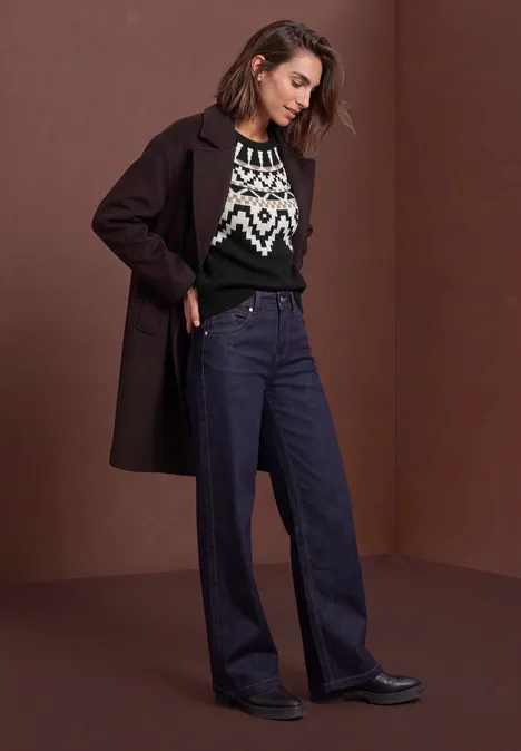 Frau in langem Mantel, gemustertem Pullover und weiten Jeans vor schlichtem braunem Hintergrund