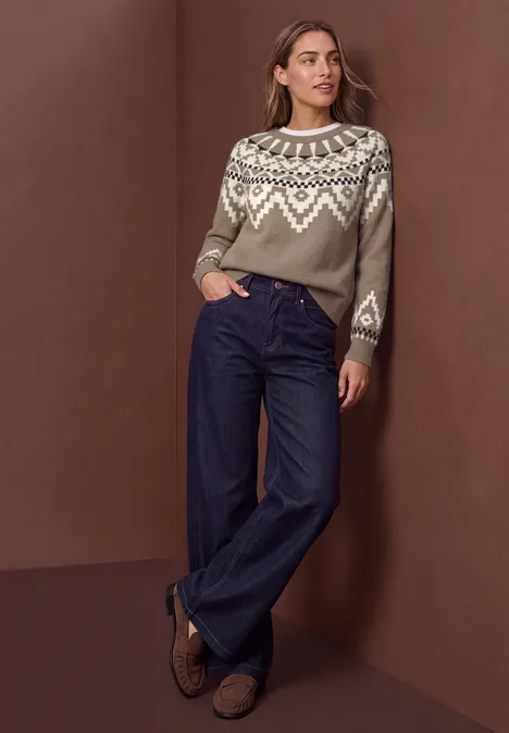 Pullover mit Rundhals und Jacquardmuster sanded amber Frau trägt gemusterten Pullover, weite Jeanshose und braune Loafers vor braunem Hintergrund.