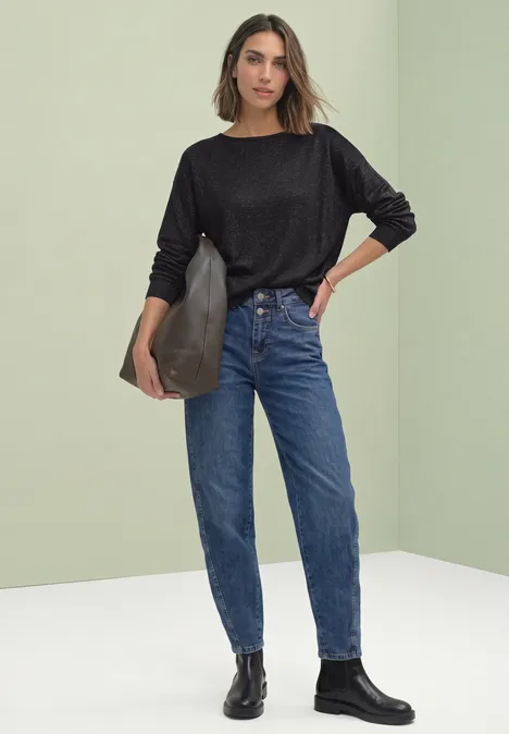 Dolman Pullover mit Glitzerdetails Black Frau in schwarzem Sweater, blauen High-Waist-Jeans, schwarzen Stiefeletten, schwarze Tasche vor hellem Hintergrund.