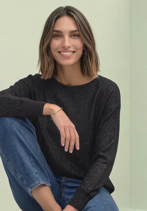 Dolman Pullover mit Glitzerdetails Black Junge Frau sitzt lächelnd in schwarzer Bluse und Jeans vor neutralem Hintergrund.