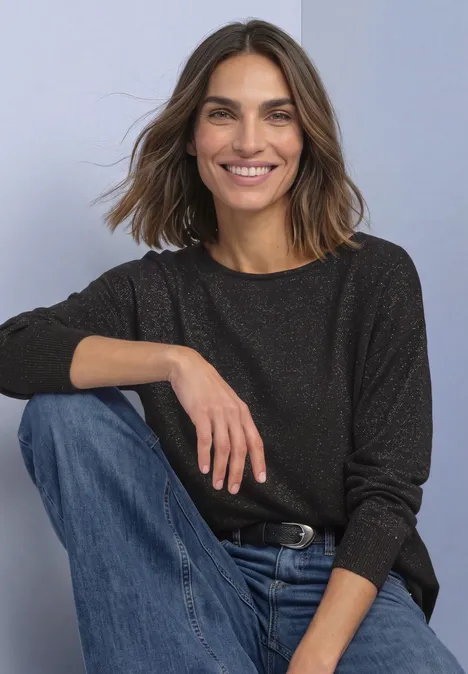 Dolman Pullover mit Glitzerdetails black coffee Eine lächelnde Frau mit braunen Haaren in dunkel meliertem Pullover und blauen Jeans sitzt entspannt.