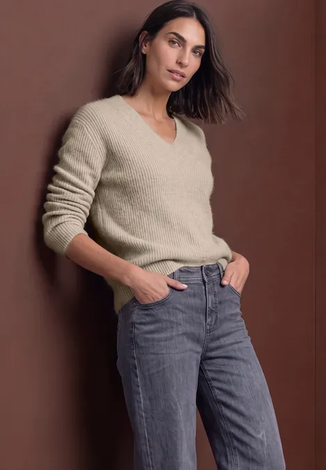 Trui van veergaren met V-hals clay sand Vrouw in een crèmekleurige V-trui en grijze jeans, ontspannen pose tegen een bruine achtergrond.