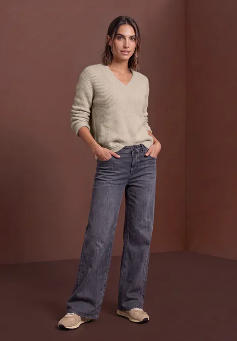 Trui van veergaren met V-hals clay sand Vrouw in beige V-pullover, wijde spijkerbroek en pantoffels, staand voor een bruine achtergrond.