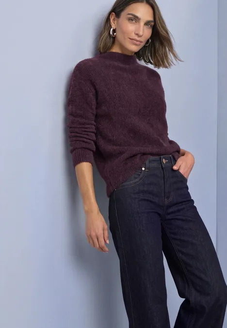 Federgarn Pullover mit Turtle Neck berry plum Frau lehnt an Wand, trägt weinroten Pullover und dunkle Jeans.