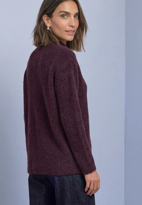 Federgarn Pullover mit Turtle Neck berry plum Frau in dunkelrotem Strickpullover und Jeans vor neutralem Hintergrund, blickt über die Schulter.