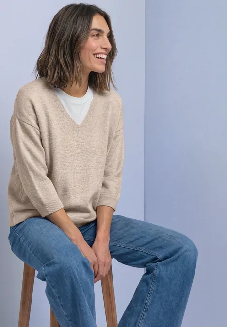 Pull à manches 3/4 et col en V clay sand mel. Eine lächelnde Frau auf einem Hocker trägt beigen V-Pullover, weißes Shirt und Jeans vor hellblauem Hintergrund