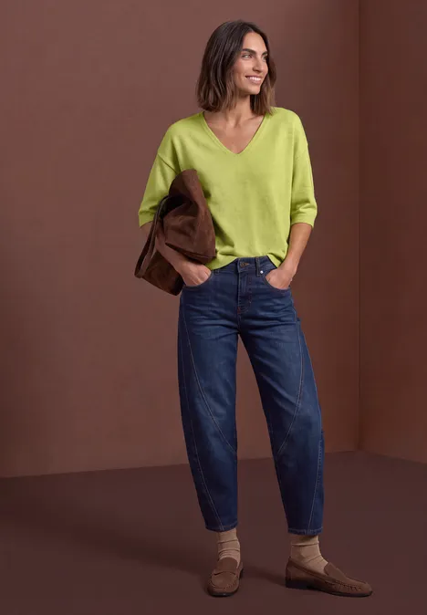 Femme en jean bleu décontracté, pull vert à col V et mocassins beiges, tenant un blazer marron.