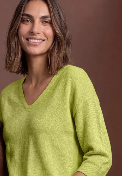 Femme souriante aux cheveux bruns, portant un pull vert clair à col V devant un fond marron.