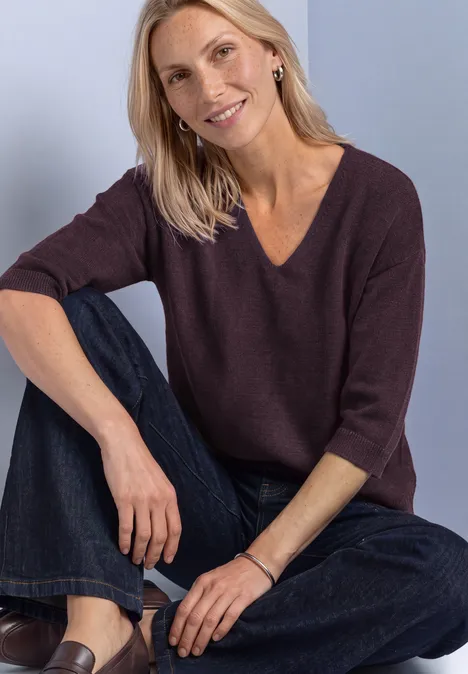 Pull à manches 3/4 et col en V berry plum Eine lächelnde Frau in lila Pullover und Jeans sitzt entspannt, Arm auf angewinkeltes Bein gestützt.